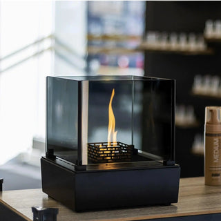 Black hexagonal table-top fireplace with flames on a wooden surface, with products in the background.