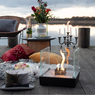 Outdoor setting with a fire pit, guitar, and pillows by a body of water.